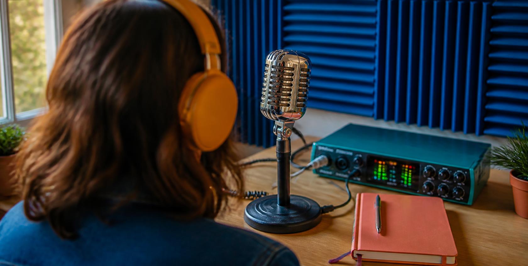 Seen from behind, a woman at a studio microphone recording an episode.
